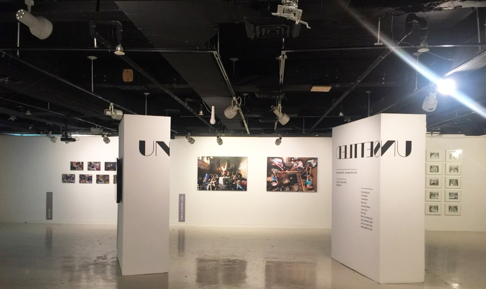 주뉴욕 한국문화원 'Unsettled' 공모전 입구에서 바라본 전시장 전경이다. 전시 제목과 전시 내용이 설명된 이동 벽 두 개와 그 뒤에 사진 작품들을 볼 수 있다.