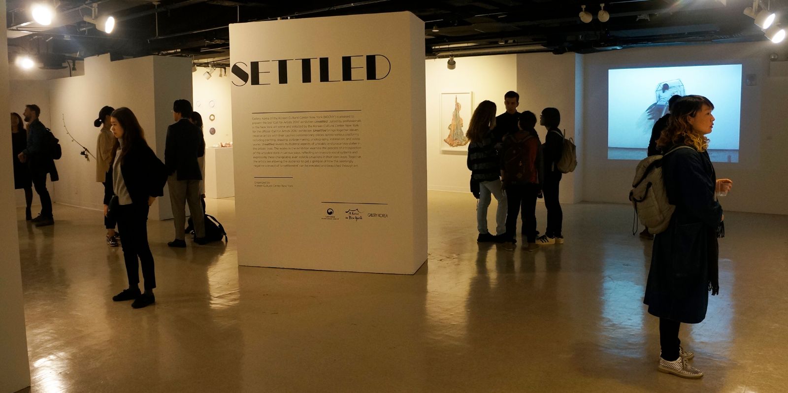 이동 벽에 'Unsettled' 공모전에 대한 전시설명을 볼 수 있다. 전시 제목의 함축적인 의미를 강조하기 위해 모서리를 이용하여 디자인한 것을 볼 수 있다. 한 관람객이 반대편에 있는 참여 작가들의 이름을 유심히 읽고 있다.